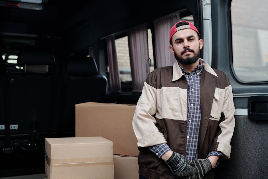 A young man with a beard and wearing a red cap, plaid shirt, and a two-tone beige and brown jacket, stands inside a van used for house removals, surrounded by cardboard boxes. The boxes are stacked be